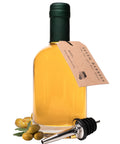 A bottle of olive oil with a dark green cap and a kraft paper hangtag reading 'SEVEN BARRELS', shown on a white background with olives and a metal pourer in front.