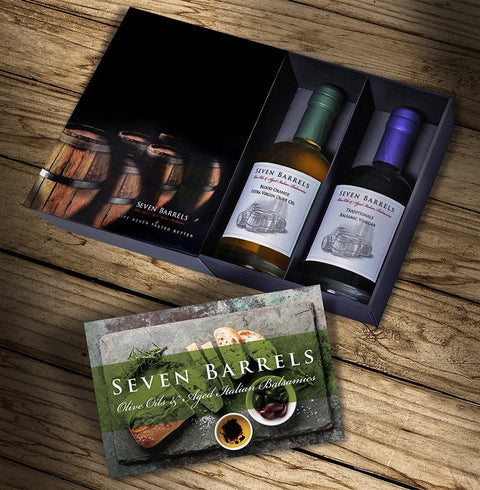 A boxed wine gift set from Seven Barrels displayed on a rustic wooden surface. The open dark presentation box contains two bottles of wine, one with a green cap and one with a purple cap, both labeled Seven Barrels.