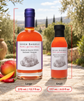 Two bottles of Seven Barrels Srubacha Mango White Balsamic Vinegar shown on a stone surface outdoors, with a large bottle labeled Large and a smaller bottle labeled Small. Red measurement lines and text indicate the sizes and volumes.